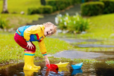 Su birikintisinde kağıt tekneyle oynayan bir çocuk. Çocuklar sonbahar yağmurunda dışarıda oynarlar. Genç çocuklar için sonbahar yağmurlu hava aktivitesi. Çamurlu su birikintilerinde atlayan çocuk. Bebek için su geçirmez ceket ve botlar..