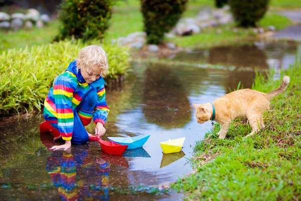 子供と猫が水たまりに紙のボートで遊んでいます 子供たちは秋の雨で屋外で遊ぶ 小さな男の子とペットの動物 子供のための秋の雨の天気屋外活動 泥だらけの水たまりに飛び込む子供 — ストック写真
