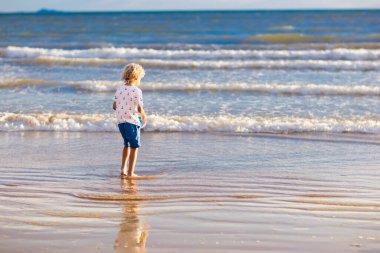 Tropik plajda oynayan bir çocuk. Deniz kıyısında elinde kova ve kürek olan küçük bir çocuk. Aile yaz tatili. Çocuk kumdan kale yapıyor. Çocuklar deniz kabuğu arar ve oynarlar. Çocuklar için su ve kum eğlencesi. 
