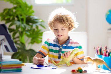 Çevrimiçi uzaktan öğrenme. Bilgisayarlı okul çocukları öğretmen ve sınıf grubuyla görüntülü konuşma yapıyorlar. Çocuk boyama kil dinozoru. Evden gelen çocuk için sanat ve el işleri. Evde eğitim.