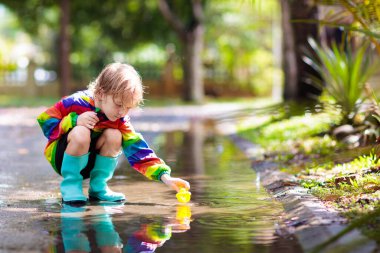 Sonbahar parkında yağmurda oynayan bir çocuk. Yağmurlu bir sonbahar gününde çamurlu su birikintisine atlayan bir çocuk. Yağmur çizmeli küçük bir çocuk ve açık havada sağanak yağmurda kırmızı ceketli. Çocuklar su geçirmez ayakkabı ve ceket giyiyor..