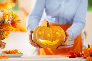 Cadılar Bayramı için balkabağı oyan bir kadın. Geleneksel şaka mı şeker mi dekorasyonu için önlüklü bir kadın Jack O feneri kesiyor. Altın sonbahar yapraklı şöminenin üzerinde çelenk olan dekore edilmiş oturma odası.