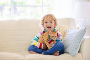 Çocuk kediyle oynuyor. Kedi yavrusu tutan çocuk. Küçük çocuk evdeki güneşli oturma odasında kanepede oturan sevimli hayvanlara sarılıyor. Çocuklar evcil hayvanlarla oynar. Çocuklar ve evcil hayvanlar.