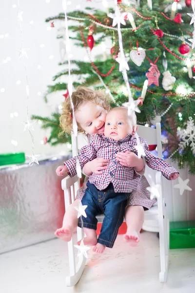 Adorable toddler girl holding her baby brother stting under a beautiful Christmas tree — Stock Photo, Image