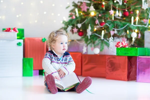 Beautiful little toddler girl reading a book sitting under a decorated new year tree — Stock Photo, Image