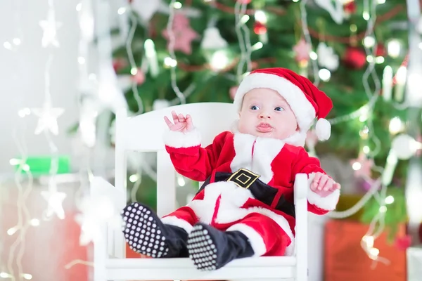 Adorable newborn baby boy in Sante outfit sitting under Christmas tree — Stock Photo, Image