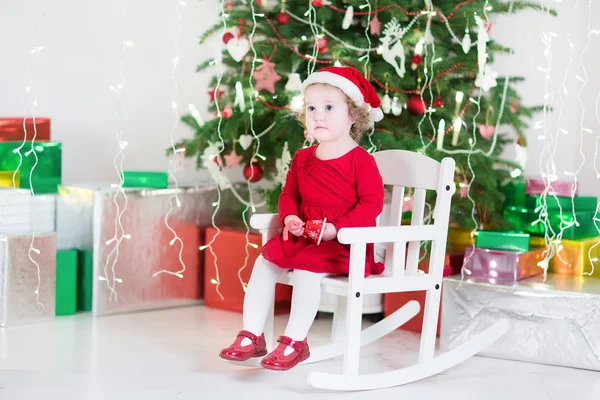 Χαριτωμένο μικρό παιδί κορίτσι σε ένα κόκκινο φόρεμα και santa καπέλο που κάθεται κάτω από το χριστουγεννιάτικο δέντρο — Φωτογραφία Αρχείου