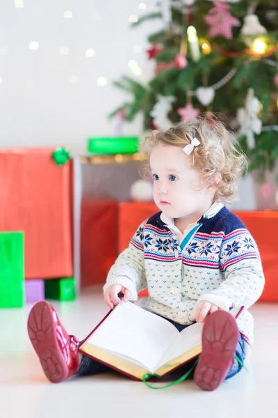 Cute toddler girl with curly hair reading a book under a beautiful tree — Stock Photo, Image