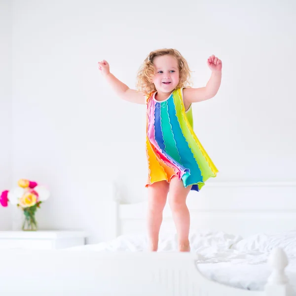 Linda niña rizada en un vestido colorido saltando sobre una gran cama blanca riendo y divirtiéndose en una soleada mañana de fin de semana en un dormitorio —  Fotos de Stock