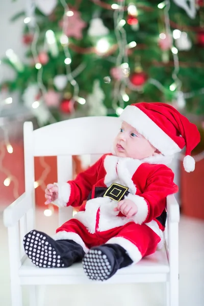 Roliga nyfödd baby boy i en santa dräkt under en dekorerad julgran — Stockfoto