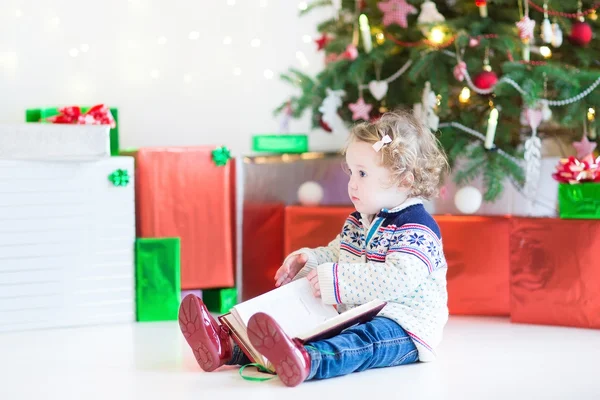 可爱的蹒跚学步的女孩在看一本书坐在圣诞树下 — 图库照片