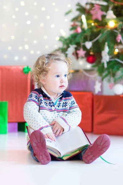 蹒跚学步的小女孩读一本书，坐在圣诞树下 — 图库照片