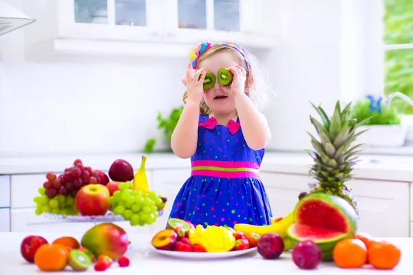 Menina encaracolado bonito em um vestido de verão colorido comendo frutas tropicais frescas e baga para lanche pequeno-almoço saudável em uma cozinha familiar ensolarada branca — Fotografia de Stock