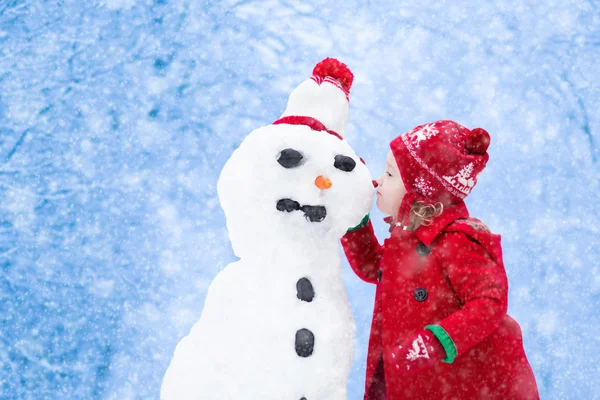 Engraçado menina criança em um chapéu nórdico de malha vermelha e casaco quente brincando com um homem de neve. As crianças brincam ao ar livre no inverno. Crianças se divertindo na época do Natal. Criança edifício boneco de neve em Xmas . — Fotografia de Stock