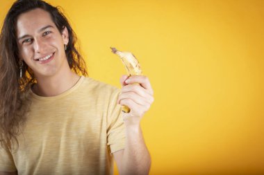 Gündelik süveter ve uzun saçlı, sarı arka planda, elinde telefon olarak muz olan genç yakışıklı bir adam.