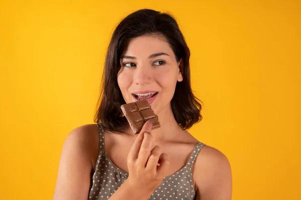 黒髪の白い女性は太ったフードチョコレートバーでカジュアルな服を着て黄色の背景で食べ物を肥やす — ストック写真