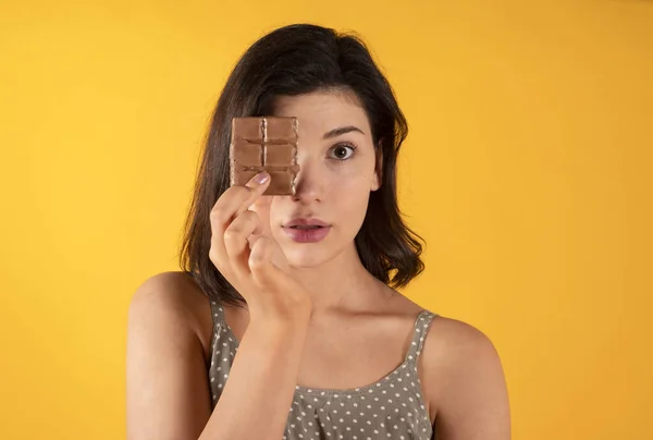 黒髪の白い女性は太ったフードチョコレートバーでカジュアルな服を着て黄色の背景で食べ物を肥やす — ストック写真