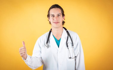 Genç erkek doktor, tamam parmakla gülümsüyor, sarı arka planda, boşluğu kopyala