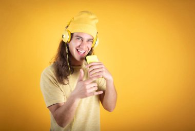 Uzun saçlı bir adam, günlük giysiler, mutlu dans, cep telefonu ve kulaklıkla şarkı söyleyen sarı arka plan, kopyalama alanı