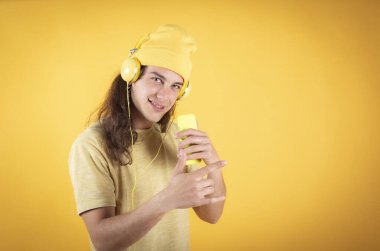 Uzun saçlı bir adam, günlük giysiler, mutlu dans, cep telefonu ve kulaklıkla şarkı söyleyen sarı arka plan, kopyalama alanı