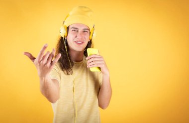 Uzun saçlı bir adam, günlük giysiler, mutlu dans, cep telefonu ve kulaklıkla şarkı söyleyen sarı arka plan, kopyalama alanı