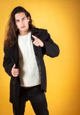 Yakışıklı, uzun saçlı, kışlık giysiler giyen, sarı arka planda parmağını gösteren, boşluğu kopyalayan bir adam.