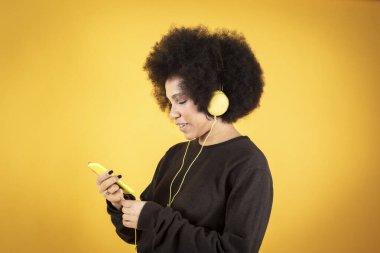 Şok edici koyu tenli bir kadının elinde telefonla oturup iyi haberler okuduğu modern kulaklıkların modaya uygun beyaz çizgili kırmızı pantolonlar giydiği sarı arka plan.