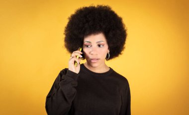 Neşeli, sevimli, kıvırcık, sarı kazaklı, kolyeli, elinde telefon olan siyah gelinin uzay fotoğrafının kopyası. Kendini parlak renkli arka plan fotokopisine karşı izole ederken neşeli, sevimli ve çekici bir kadının uzay fotoğrafçılığına karşı.