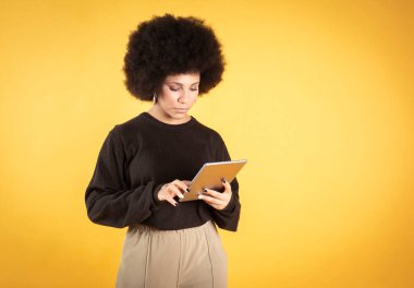 Güzel bir afro kadının sarı arka planda tabletini kullanırken fotoğrafını çek, boşluğu kopyala
