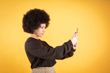 Karışık afro kadın, akıllı telefonunu kullanıyor, mutlu, selfie, sarı arka plan, kopyalama alanı