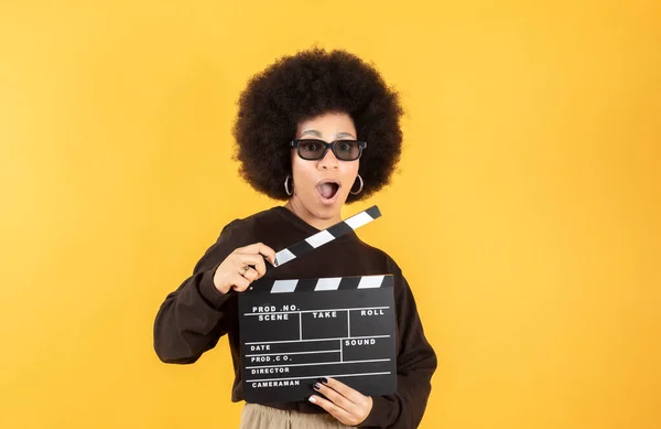Relaxed, mixed afro beautiful young woman in eyeglasses holds classic black film making clapperboard isolated on yellow background. Sincere people emotions, lifestyle concept. Advertising area