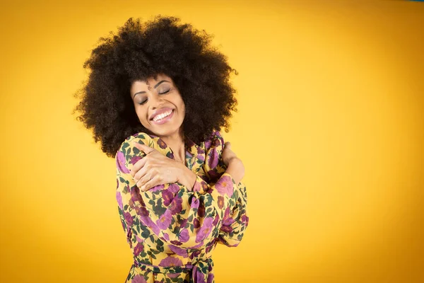 Mutlu yüz ifadesini tatmin eden, gözlerini kapayan, gülümseyen, kendini kucaklayan, sevgi ve minnettarlıkla dolu zarif mavi pantolonlu Afro-Amerikan bir kadının stüdyo fotoğrafı.