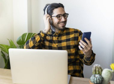 Gözlüklü yakışıklı bir adam bilgisayarın önünde oturmuş müzik dinlemek için cep telefonunu kullanıyor.