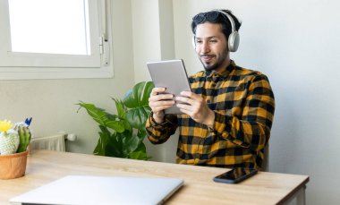 Adam ofiste tablet kullanarak oturuyor ve masanın üstünde de başka bir dizüstü bilgisayar var.