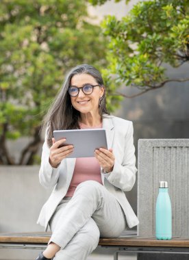 Elinde tablet ve bankta meşrubat olan olgun bir iş kadını.