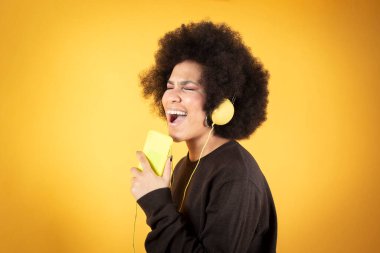 Güzel afro kadın kulaklık, akıllı telefon ve sarı arka planda müzik dinliyor.