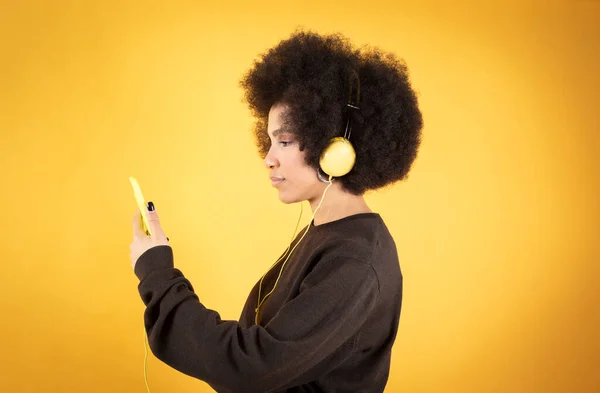 漂亮的非洲女人 带着耳机和智能手机听音乐 背景是黄色的 — 图库照片