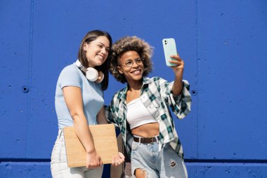 İki çok ırklı arkadaş, güzel mavi bir duvarda, kulaklık ve dizüstü bilgisayarla akıllı dille fotoğraf çekiyorlar.