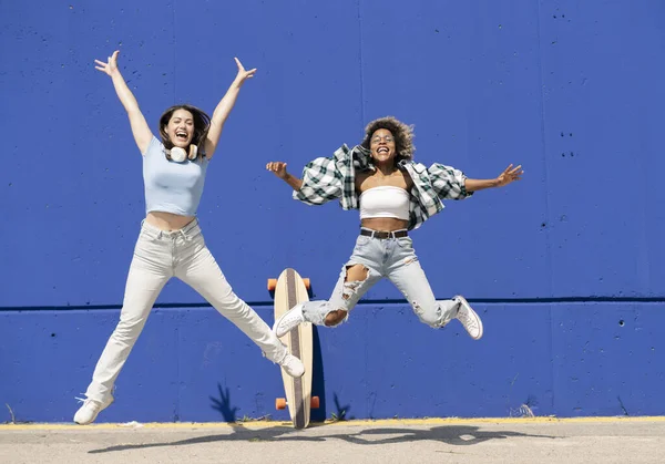 Dwie Wielonarodowe Kobiety Skaczące Niebieskiej Ścianie Deskorolką Ubraniami Casual — Zdjęcie stockowe