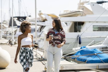 Kadınlar kahve içerek konuşuyor. Marinada poz veren iki kadın, arka planda yatlar. Kızlar deniz teknesine gidiyor.