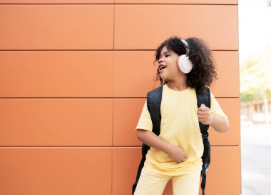 Küçük afro çocuk turuncu bir arka planda duruyor ve kulaklıkla çok mutlu müzik dinliyor.