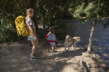 Sırt çantalı, çocuklu ve tasmalı bir kadın akan bir nehrin kenarında gölgeli toprak patikada yürüyor, yeşil orman doğasında güneşli bir aile gezisinin tadını çıkarıyor.