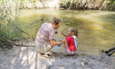 Büyükanne ve torun nehir kıyısında bir bağ anının keyfini çıkarıyorlar, şişeleri tutup etkileşime geçiyorlar. Doğada oynuyorlar, mutlu, nesilsel bir deneyimi paylaşıyorlar.