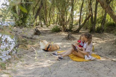 Anne ve kızı, bir nehrin kenarındaki bir battaniyede birlikte kitap okuyarak birbirlerine bağlanıyorlar. Köpekleri kumun üzerinde rahatça uzanıyor, dışarıda ailecek vakit geçiriyorlar.