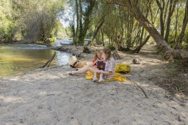 Anne ve küçük çocuk bir nehrin kenarında kumların üzerinde oturup birlikte kitap okurken köpekleri yanlarında dinleniyor ve doğada huzurlu bir günün tadını çıkarıyorlar.