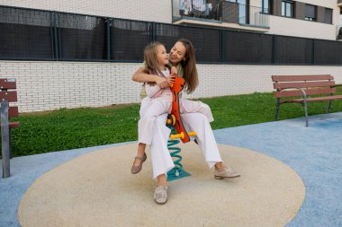 Anne ve kızı, modern bir konut parkında bir bahar binicisine gülümseyip kucaklaşıyorlar. Neşeli, neşeli bir bağ kurma anını ve tasasız çocukluk sevincini paylaşıyorlar.