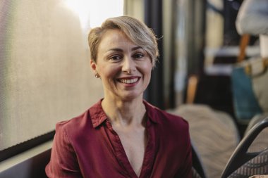 Peri tıraşlı olgun bir kadın otobüs penceresinin kenarında oturur, yol boyunca gülümser ve rahatlar, sessiz seyehat zamanının tadını çıkarır ve kameraya bakar.