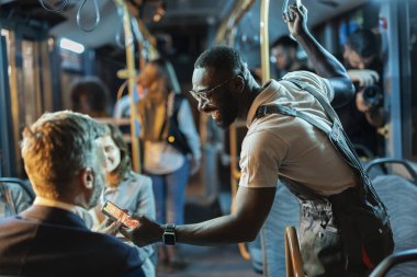 Genç bir adam, bir başka yolcuya akıllı telefondan içeriğini gösteriyor. Kalabalık bir otobüste ayakta durup gülümserken, gece ulaşımı sırasında, teknolojiyi ve bağlantıyı paylaşırken.