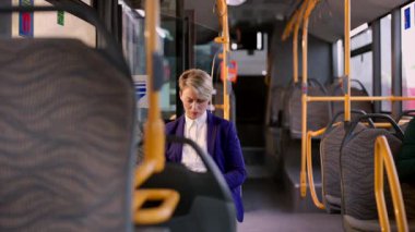 Kısa sarı saçlı profesyonel bir kadın şık bir mor takım elbise giyiyor, neredeyse boş bir toplu taşıma otobüsünün içinde duruyor, muhtemelen akıllı telefonuna ya da biletine bakıyor.