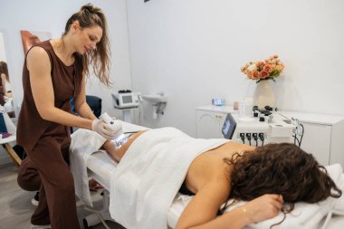 Güzellik uzmanı, profesyonel bir sağlık kliniğinde ileri teknolojiyi kullanarak bir hastanın bacağına estetik işlem uygulayıp cilt bakımı ve vücut sağlığı üzerine yoğunlaşıyor.